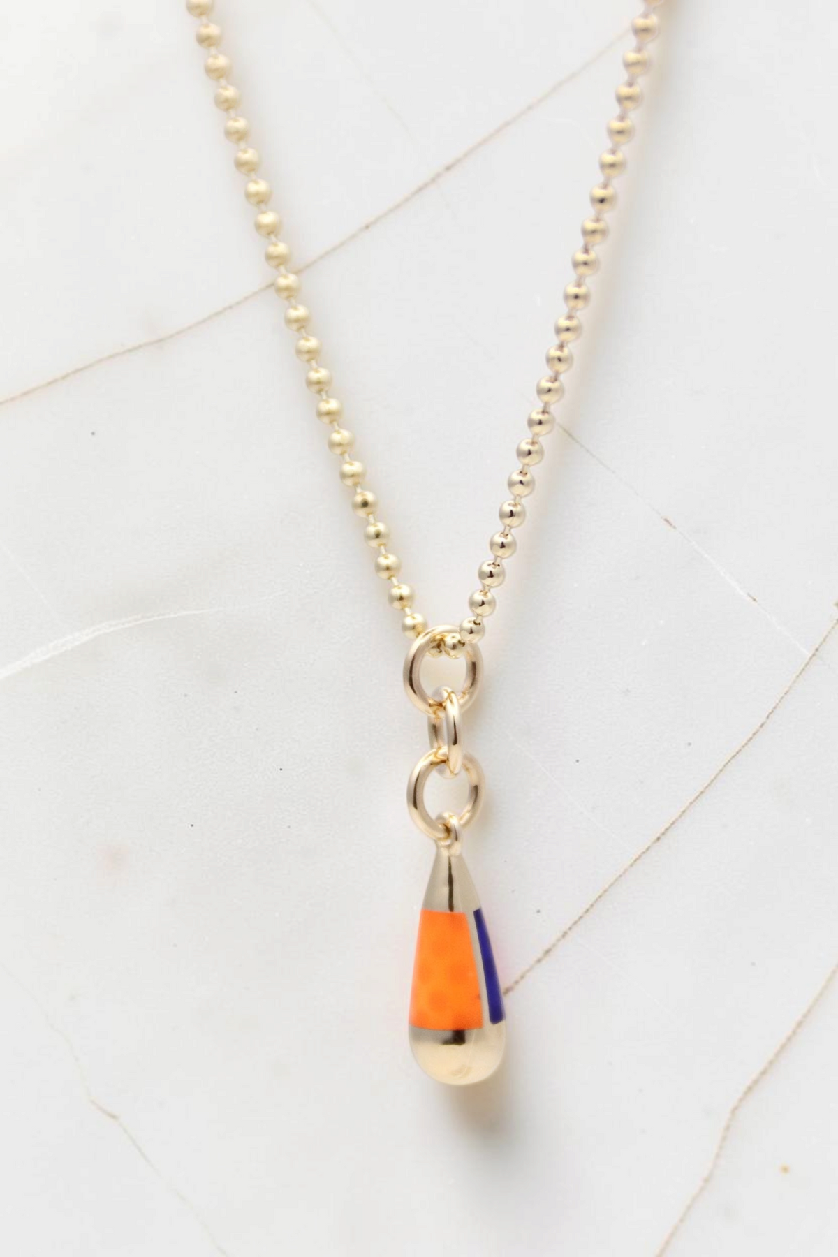 Zafrah 14k gold beaded chain necklace featuring an orange and blue enamel drop pendant on a white marble background.