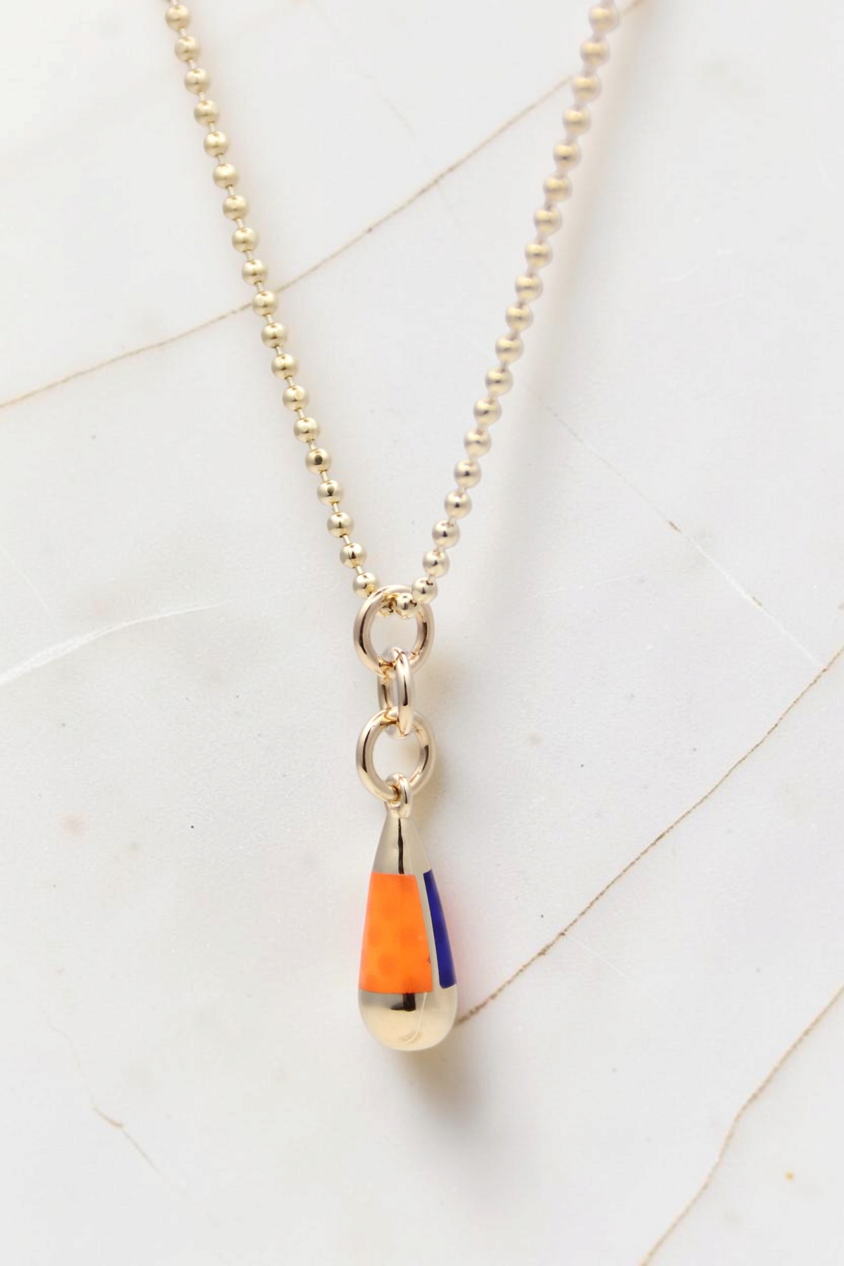 Zafrah 14k gold beaded chain necklace featuring an orange and blue enamel drop pendant on a white marble background